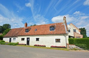 Ein zweistöckiges Steinhaus mit einem Ziegeldach, ein kleiner Garten mit roten Blumen und einem strahlend blauen Himmel.