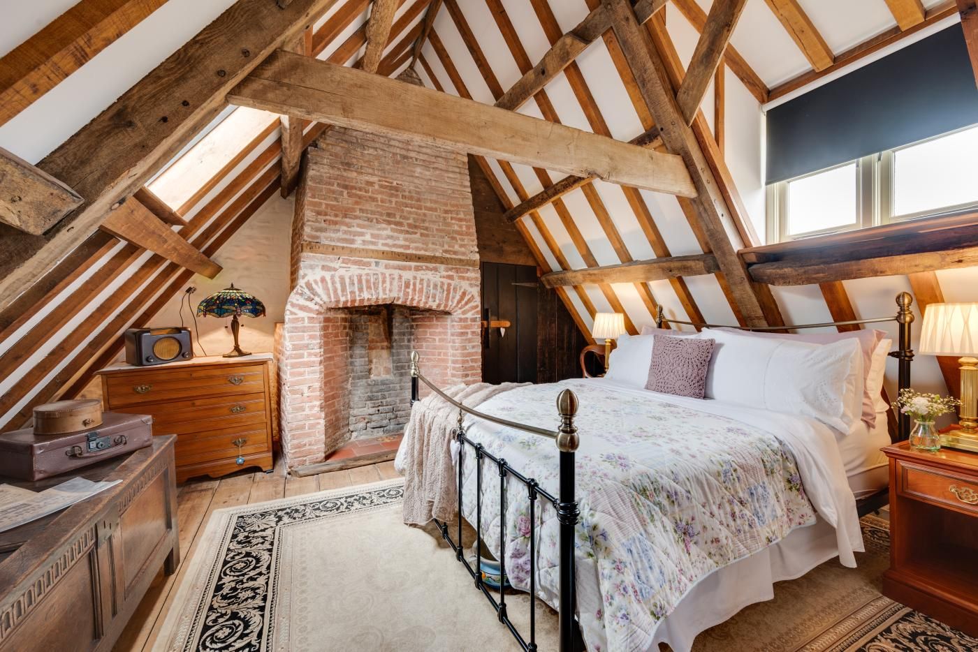 A cozy attic bedroom with exposed wooden beams, a brick fireplace, a bed with floral bedding, and a vintage radio.