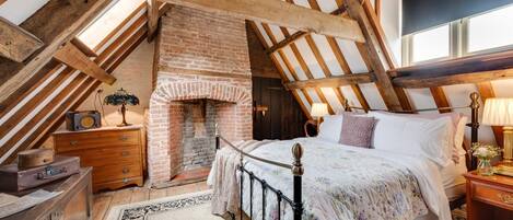 A cozy attic bedroom with exposed wooden beams, a brick fireplace, a bed with floral bedding, and a vintage radio.