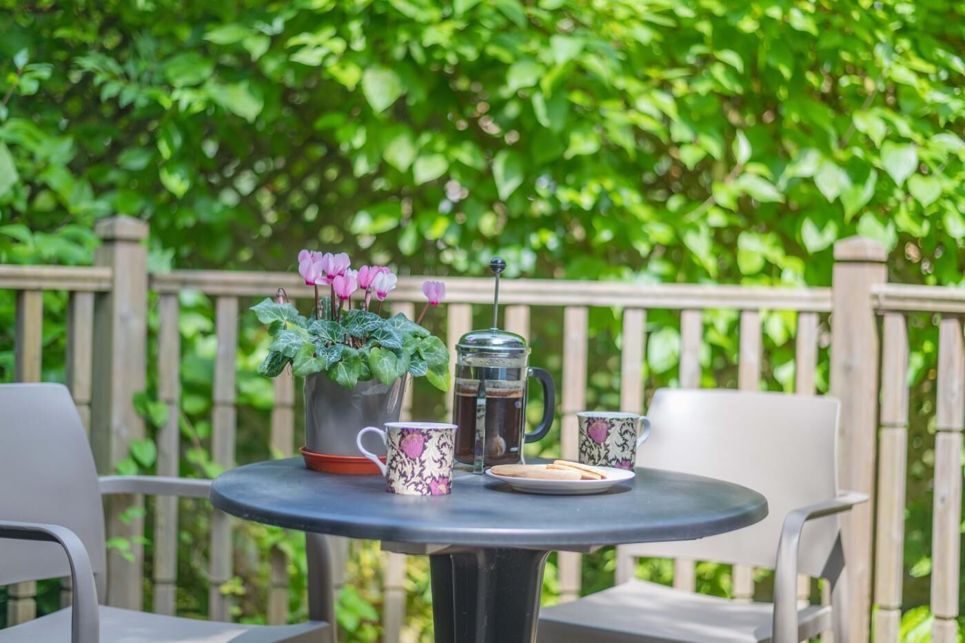 A round table with a coffee pot, two patterned cups, a plate with cookies, and a potted plant with pink flowers.