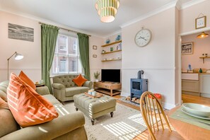 A living room with a sofa, armchair, and ottoman, a television, a fireplace, and a kitchen area with a sink and shelves. - 2 Bed in Cromer (oc-s32971) (Cromer)