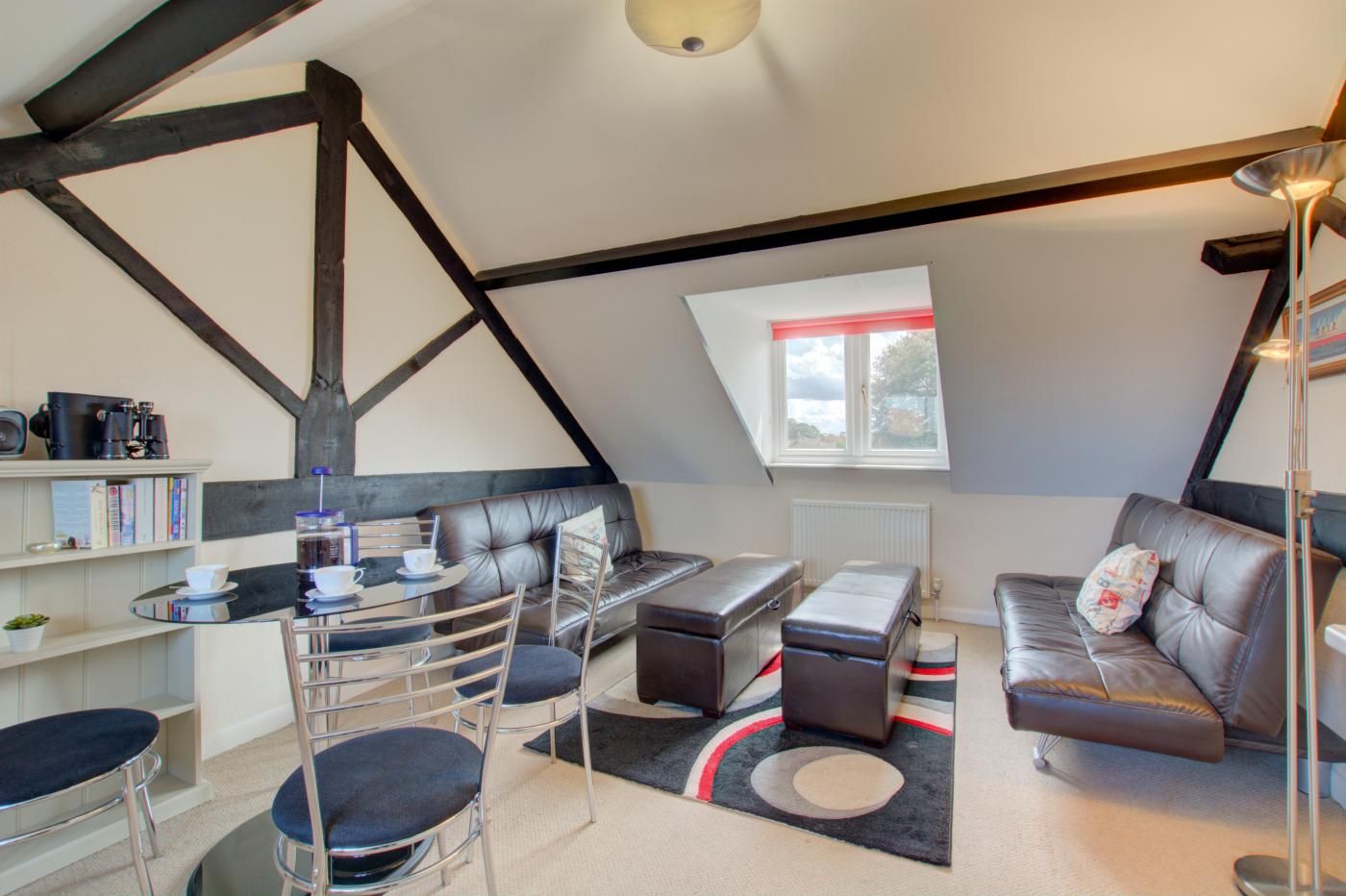 A loft-style room with exposed beams, a dining area with a round table and chairs, a sofa, and a window with a view.