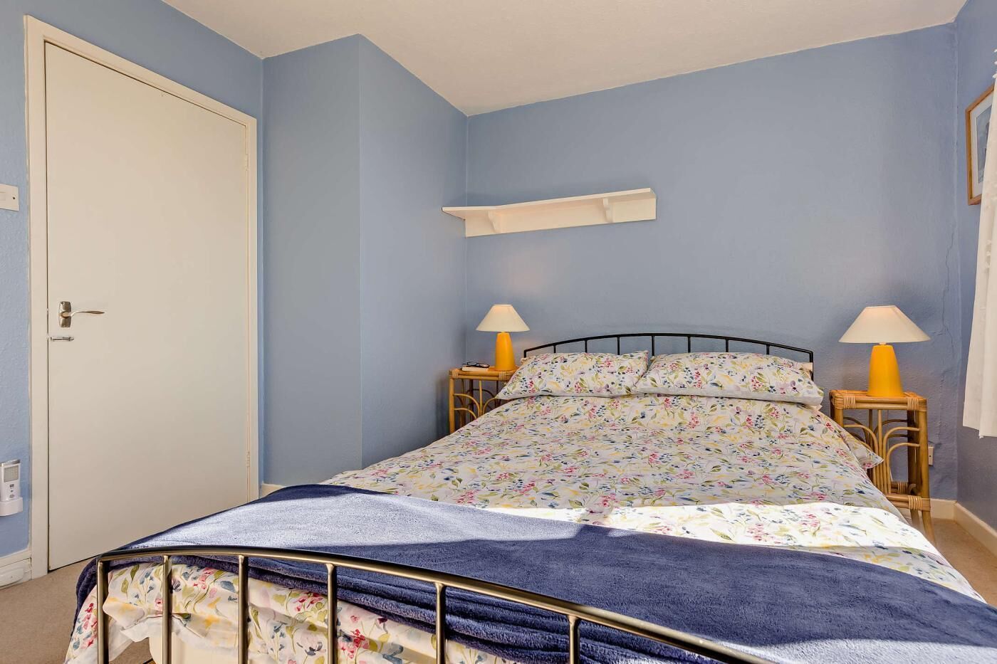 Een slaapkamer met een bed, twee bedlampjes, een witte deur en een klein plankje boven het bed.