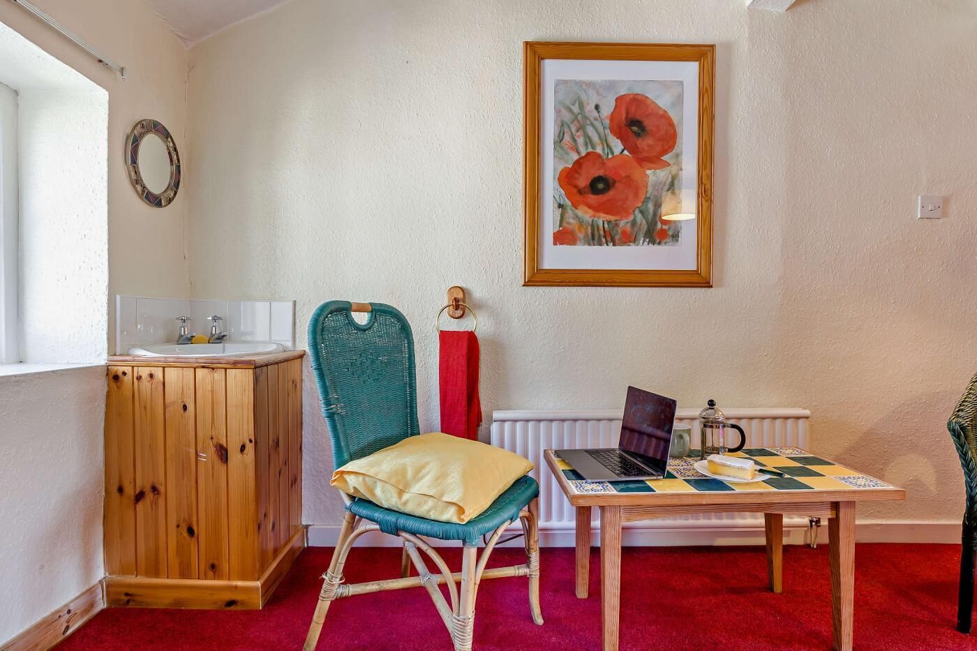 A room with a wooden cabinet, a wicker chair, a table with a laptop, and a framed painting on the wall.