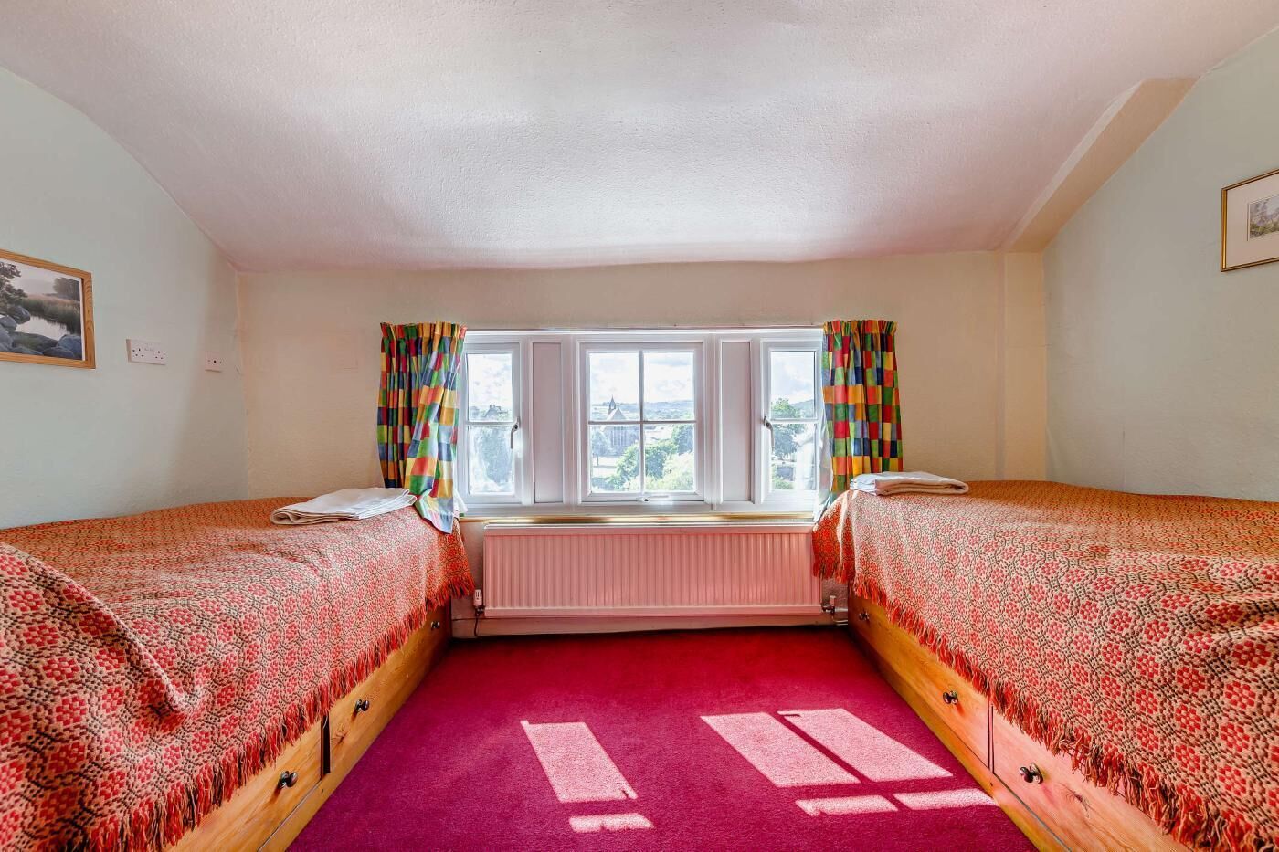 A room with two beds, a window with curtains, a radiator, and a view of the outside.