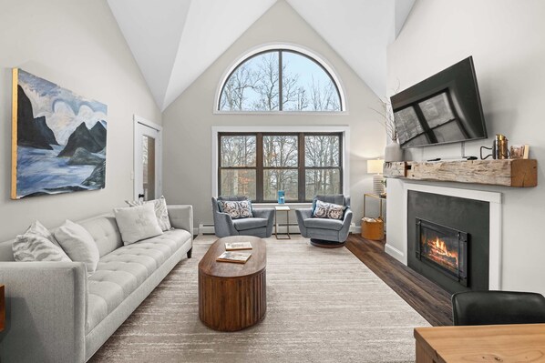 A spacious living room with a high ceiling, a fireplace, a large TV, and a cozy seating area with a sofa and two armchairs. - Luxury Winterplace Condo Steps from the Lifts (Ludlow)