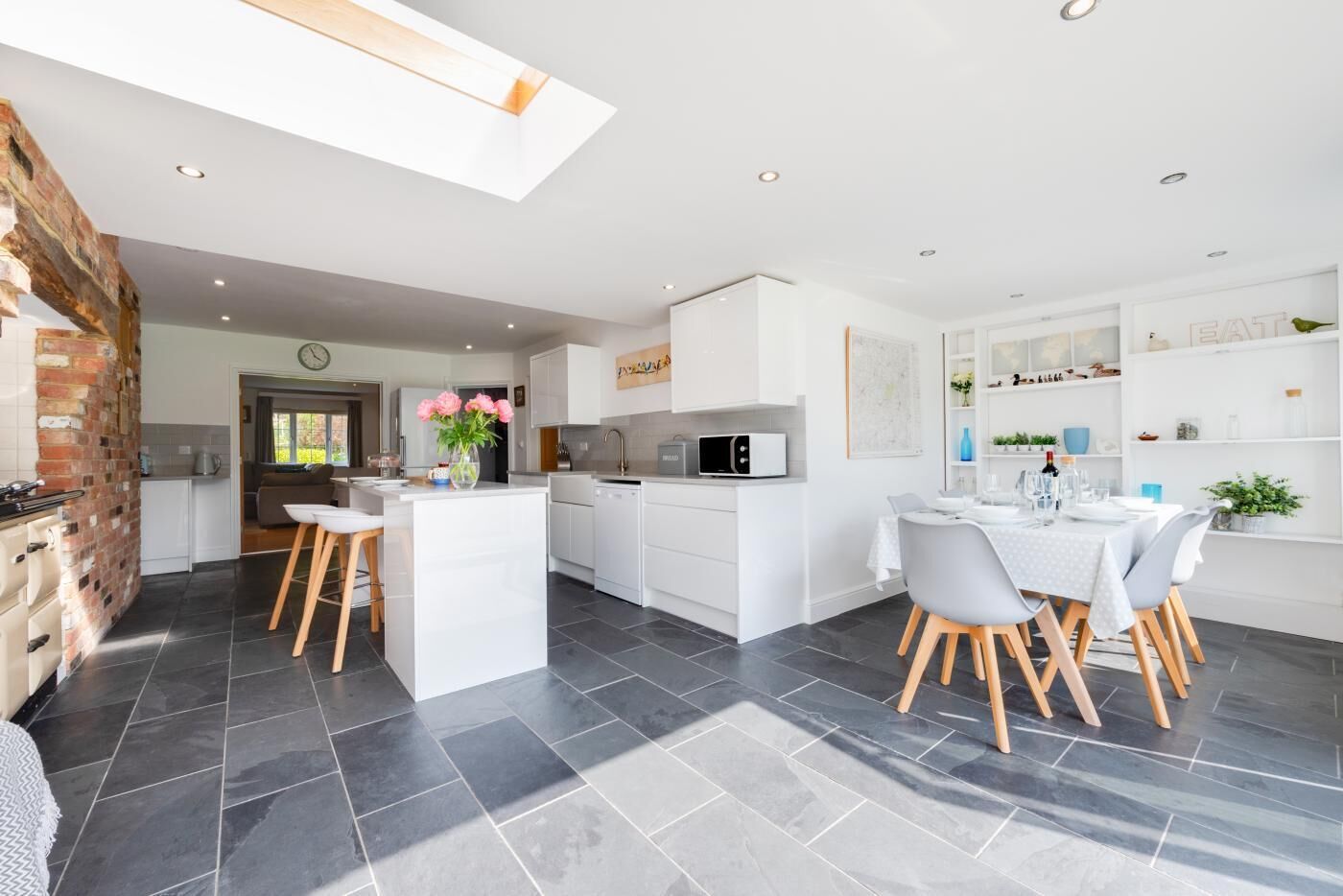 A modern kitchen with white cabinetry, a dining area with a white table and grey chairs, and a brick wall on one side.