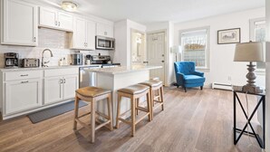 A modern kitchen with white cabinets, a central island with stools, a blue armchair, and a lamp. - Bright 2BR Escape 2 Min to Okemo, Walk to Downtown (Ludlow)