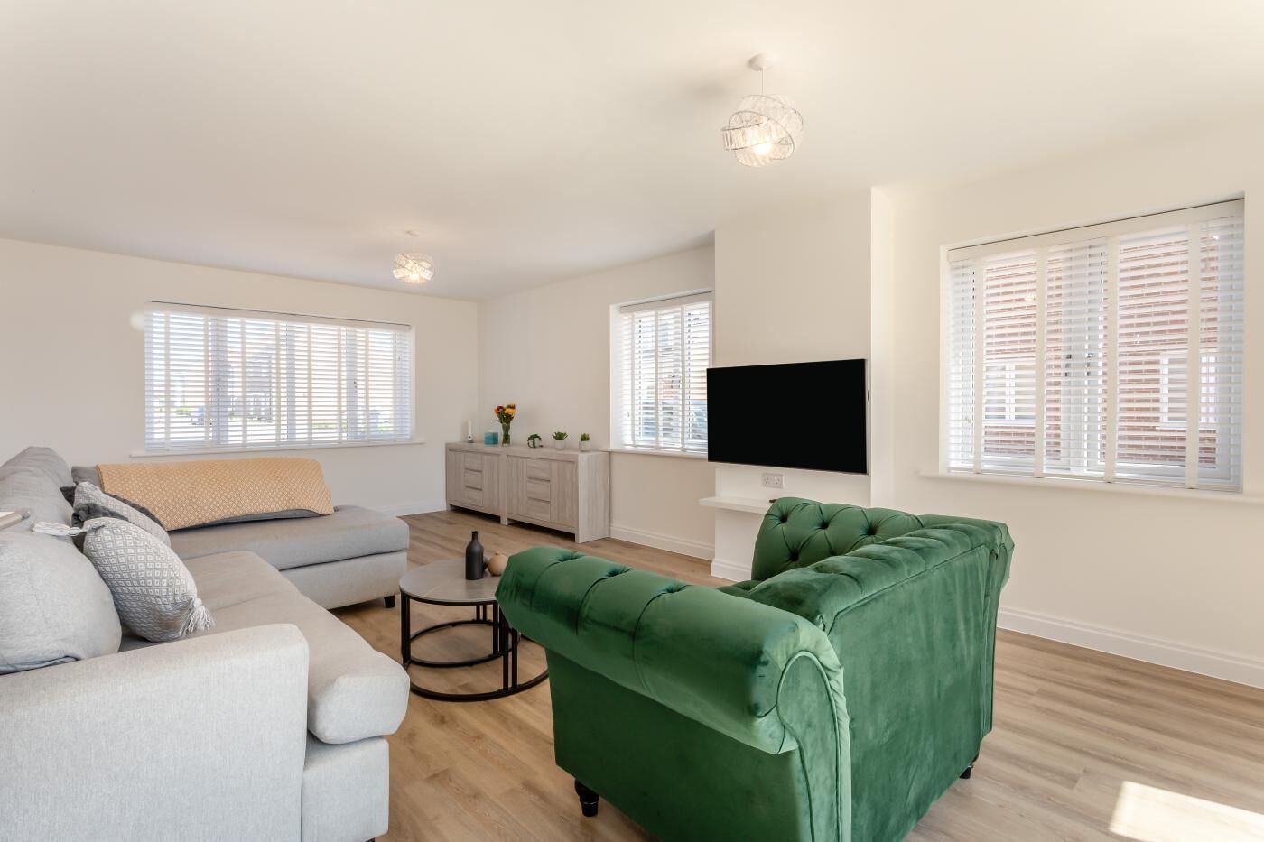 Een moderne woonkamer met een grijze bank, een groene fauteuil, een ronde salontafel en een flatscreen televisie aan de muur.
