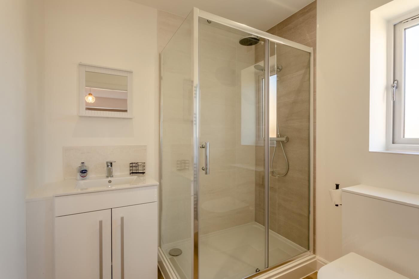 A modern bathroom with a glass-enclosed shower, a white vanity with a mirror, and a window above the sink.