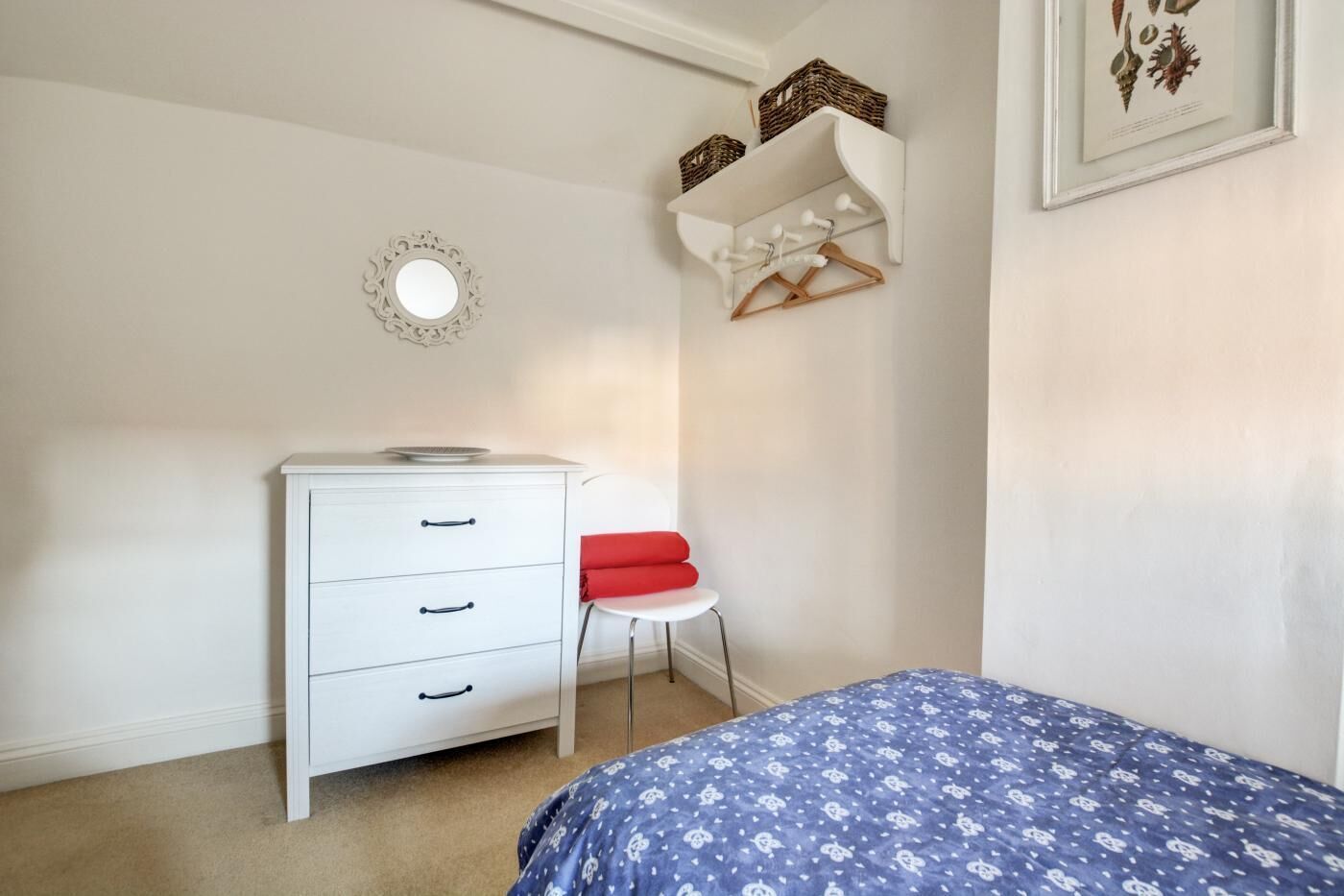 Une chambre avec une commode blanche, une chaise rouge, une couvre-lit bleue, une étagère fixée au mur et un tableau encadré.