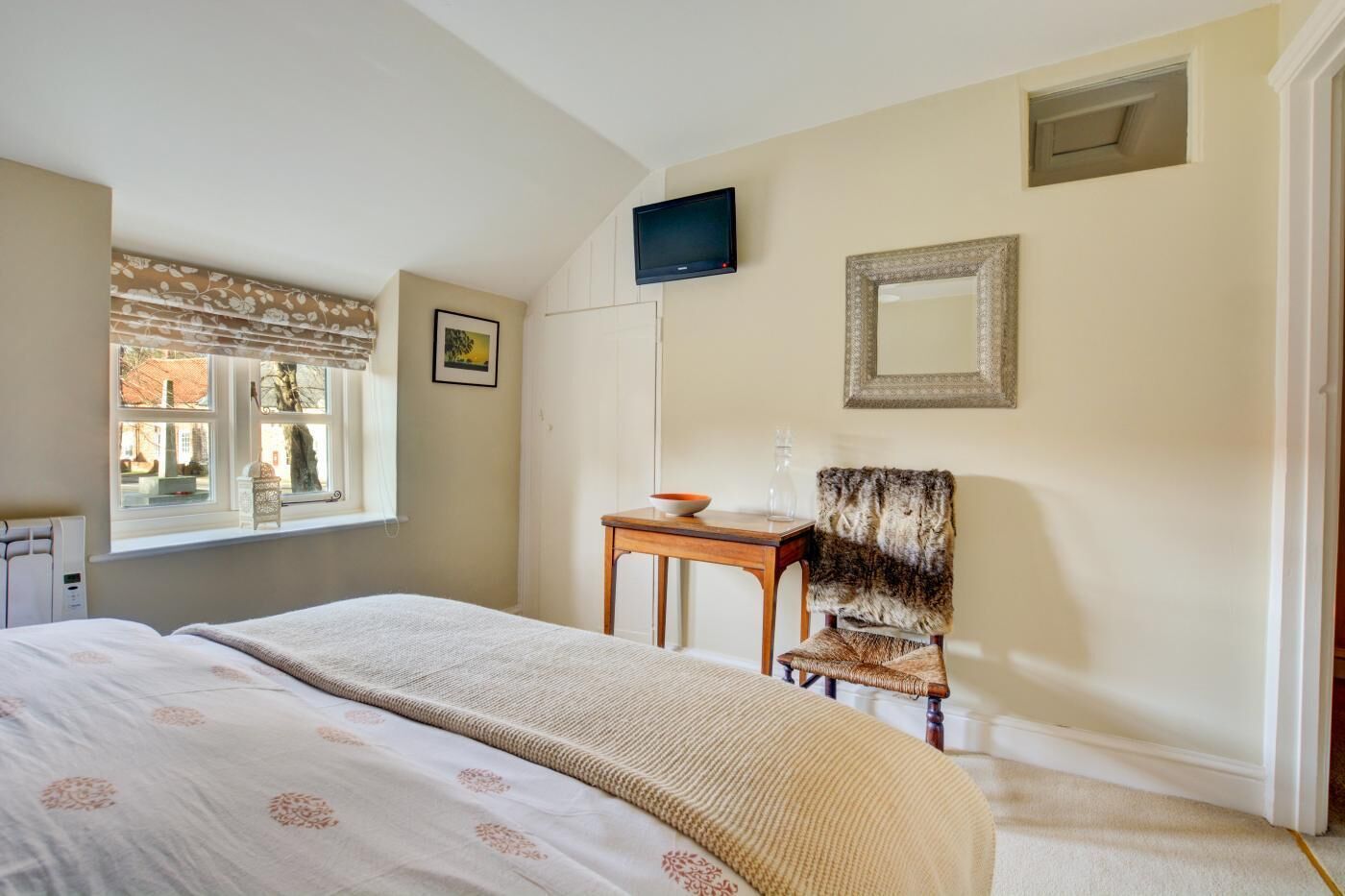 A bedroom with a bed, a chair, a table, a TV, a window, and a mirror.