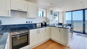 A modern kitchen with white cabinets, a black countertop, and an oven. There is a dining area with a table and chairs, and a large window offering a view of the ocean. - 2 Bed in Sheringham (oc-1540) (Sheringham)