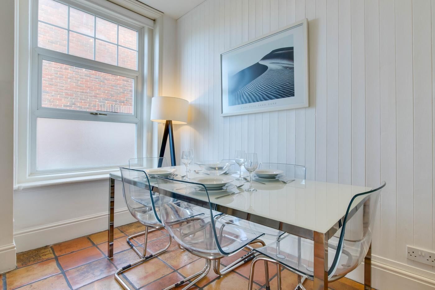 A dining area with a glass table and chairs, a framed picture on the wall, and a window with a view of a brick building.
