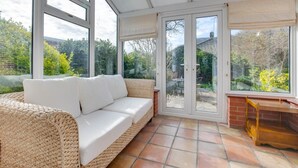 Une pièce baignée de lumière, avec un canapé en osier, une table en bois et une vue sur un jardin, visible à travers de grandes portes vitrées.