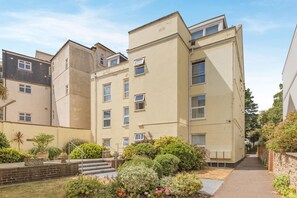 A multi-story residential building with a central staircase, a small garden area with various plants, and a clear sky. - 2 Bed in Sidmouth (oc-t32948) (Sidmouth)