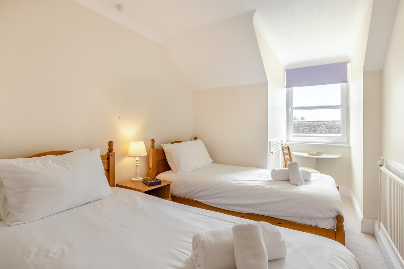 Une chambre d’hôtel avec deux lits, une table de chevet avec une lampe, une petite table avec une chaise et une fenêtre donnant sur un paysage agréable.