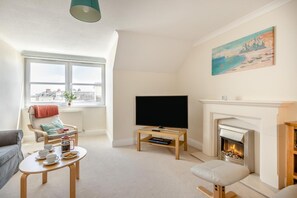 A living room with a fireplace, a television, and a window with a view of buildings. - 2 Bed in Sidmouth (oc-t32948) (Sidmouth)