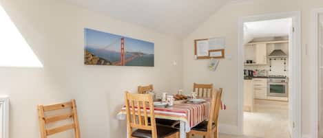 A dining area with a table set for four, a kitchen visible through an open door, and a painting of a bridge on the wall.