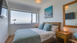 Une chambre avec une grande fenêtre, un lit avec une literie bleue et blanche, une table de chevet en bois, une lampe de chevet et un tableau accroché au mur.