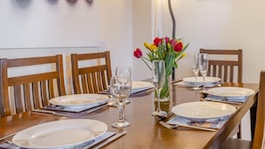Mesa de jantar posta para quatro pessoas, com cadeiras de madeira, um vaso com flores e um quadro na parede.