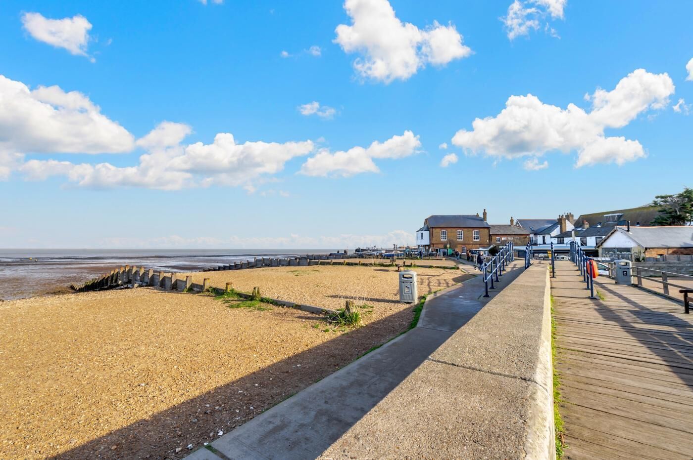 A coastal walkway with a wooden boardwalk, a sandy beach, and a building with a brick facade.