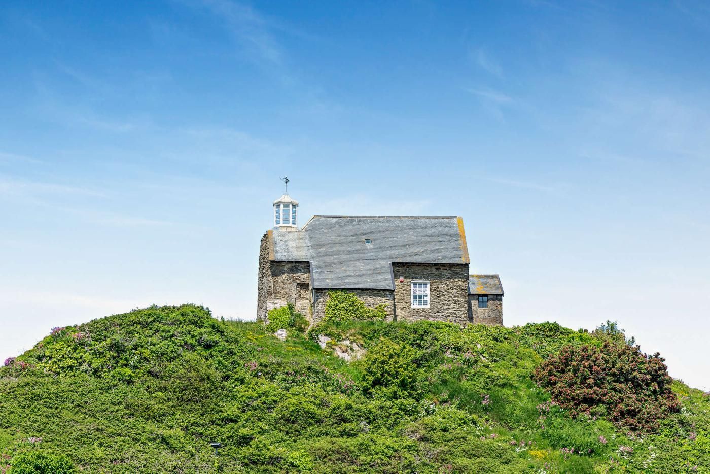 A stone building with a tower on a hill, surrounded by greenery.