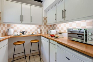 A modern kitchen with white cabinets, a microwave, a toaster, and a coffee maker. There are two stools and a dishwasher. - 2 Bed in Ilfracombe (oc-quay) (Ilfracombe)