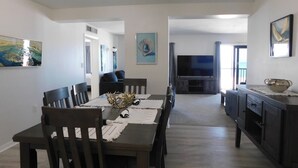 Uma sala de jantar com mesa e cadeiras de madeira escura, uma TV ao fundo e obras de arte nas paredes.