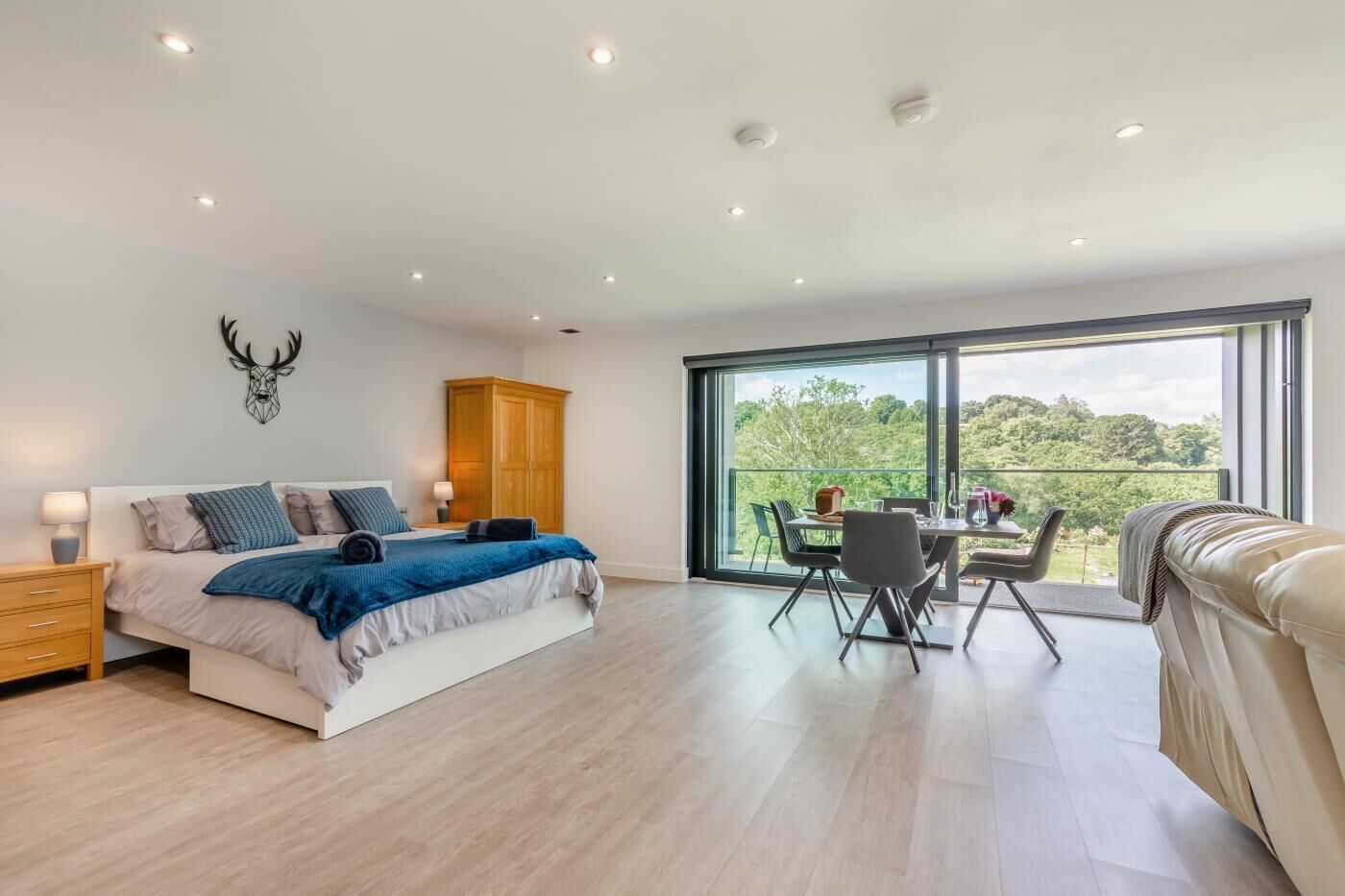 A modern bedroom with a large bed, a wooden wardrobe, a dining table with chairs, and a balcony with a view of greenery.