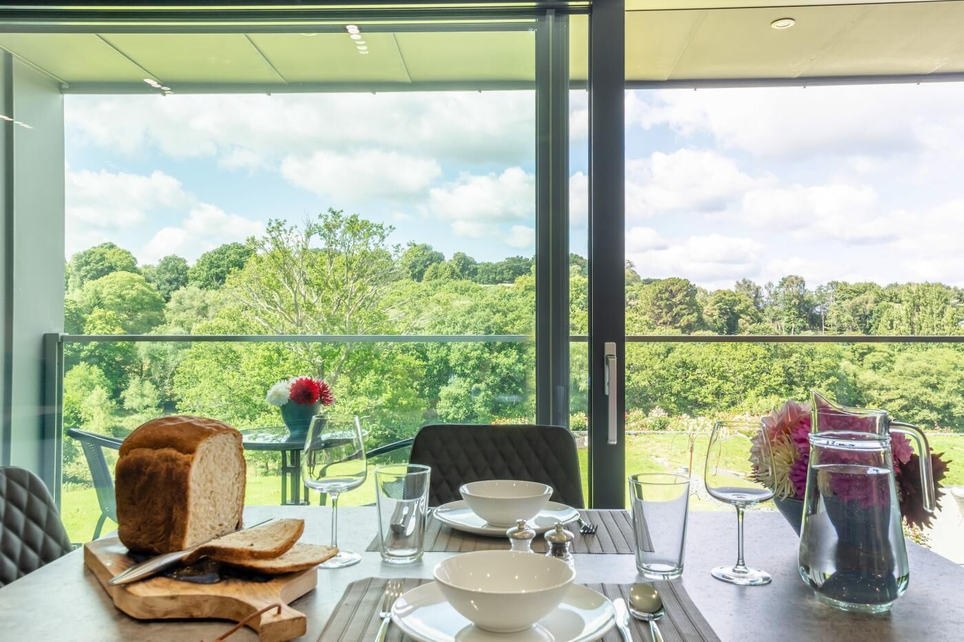 Ein Esstisch mit Brot, Weingläsern und einer Karaffe, vor einer malerischen Aussicht auf eine üppige Landschaft.