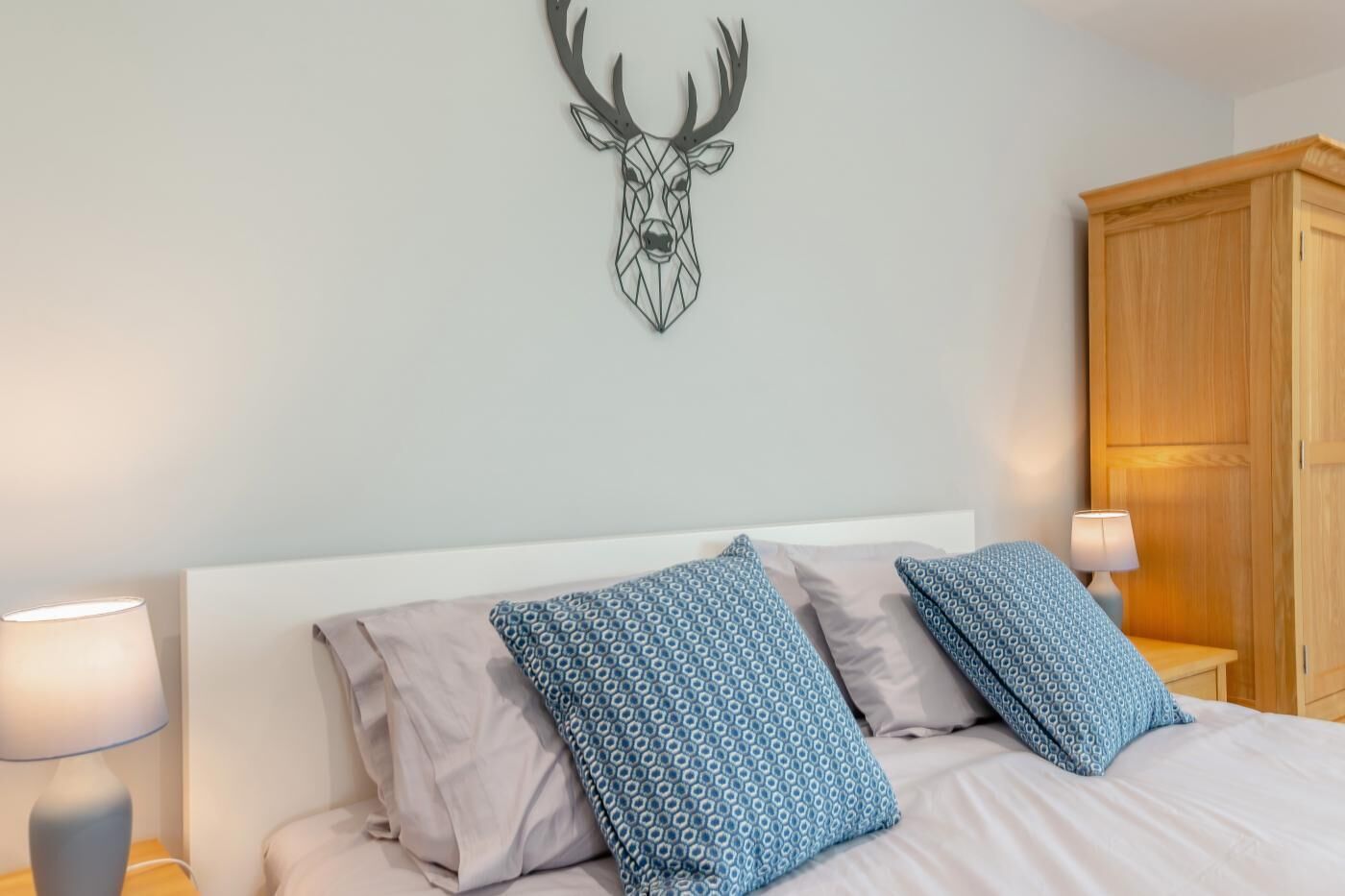 Une chambre à coucher avec un lit, deux coussins à motifs, une décoration murale représentant une tête de cerf, une armoire en bois et des lampes de chevet.