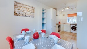 Een praktische keuken met witte kasten, een wasmachine en een eethoek met een tafelkleed met stippen en rode stoelen.
