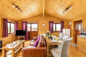 A cozy cabin interior with a wooden ceiling, a dining area with a table and chairs, a leather sofa, a television, and a kitchen area.