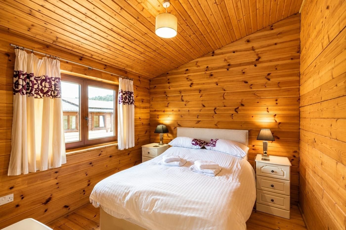 A wooden cabin bedroom with a bed, two bedside tables, a window with curtains, and a ceiling light.