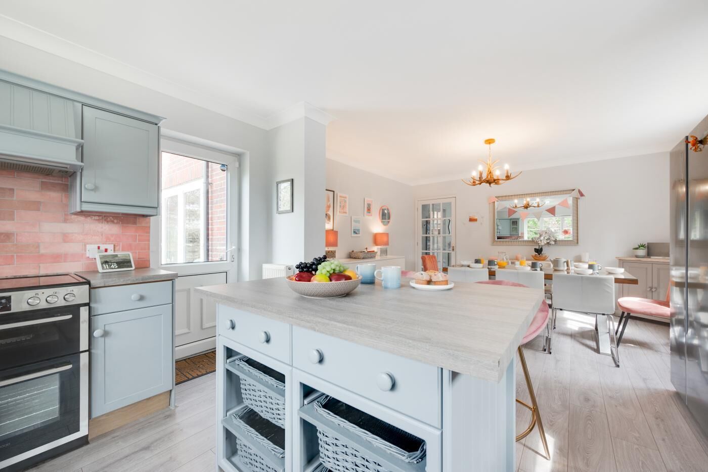 A modern kitchen with light blue cabinets, a large island with a fruit bowl, and a dining area with a chandelier.