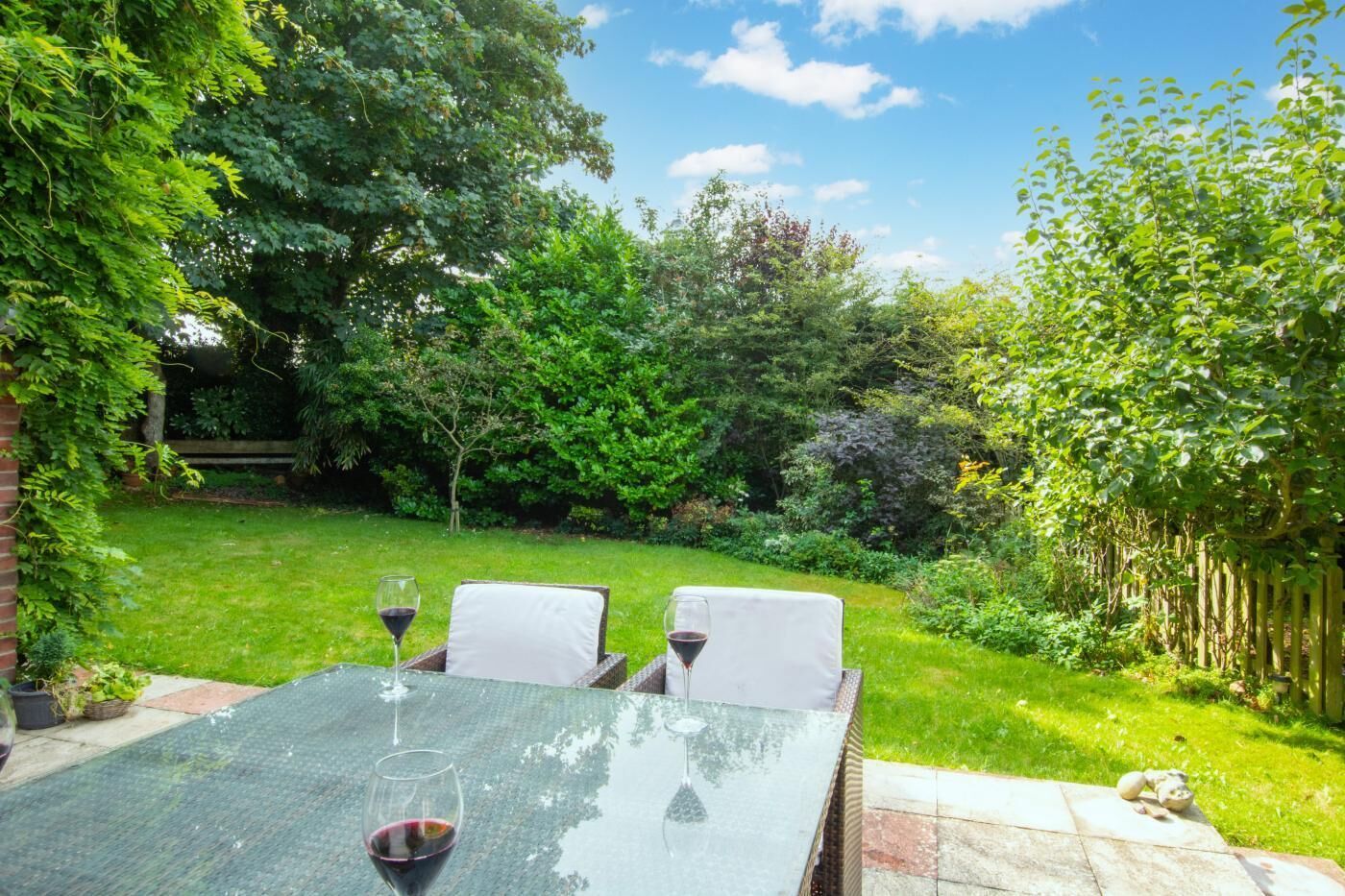 Une table de patio avec deux chaises et deux verres à vin, entourée d’une végétation luxuriante et d’une clôture en bois.