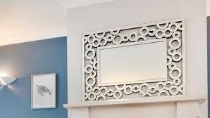 Un salon avec une cheminée, un mur bleu, un fauteuil bleu clair, une table basse blanche avec des tasses et des pâtisseries, et un miroir décoratif au-dessus de la cheminée.