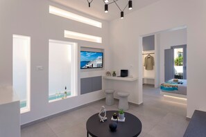 A modern hotel room with a large window, a flat-screen TV, a round table with decorative items, and a bed with a blue and white patterned bedspread. - LInea Luxury Suites (Fira)