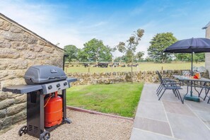 Un espace extérieur aménagé en terrasse, avec un barbecue, un mur en pierre et une vue sur un champ verdoyant où paissent des vaches.