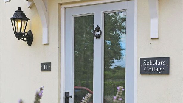 A white door with a glass panel, a black doorbell, and a sign reading "Scholars Cottage".