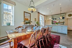 Une salle à manger avec une table en bois dressée pour six personnes, entourée de chaises. On aperçoit, au fond, une cuisine avec des placards bleu clair et une fenêtre.