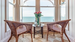 Ein Zimmer mit zwei Korbstühlen, die auf ein großes Fenster mit Meerblick ausgerichtet sind. In der Mitte steht ein kleiner Tisch mit einem Blumenstrauß und einer Kaffeekanne.