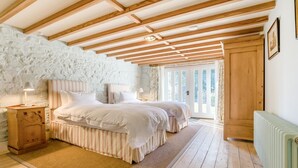 Une chambre avec un mur en pierre, des poutres apparentes, un lit double, des tables de chevet et une vue sur l’extérieur, visible à travers une porte.