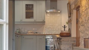 A kitchen with light grey cabinets, a stone wall, and a wooden staircase leading to a dining area. - 3 Bed in Hardraw (oc-ds1029) (Appersett)