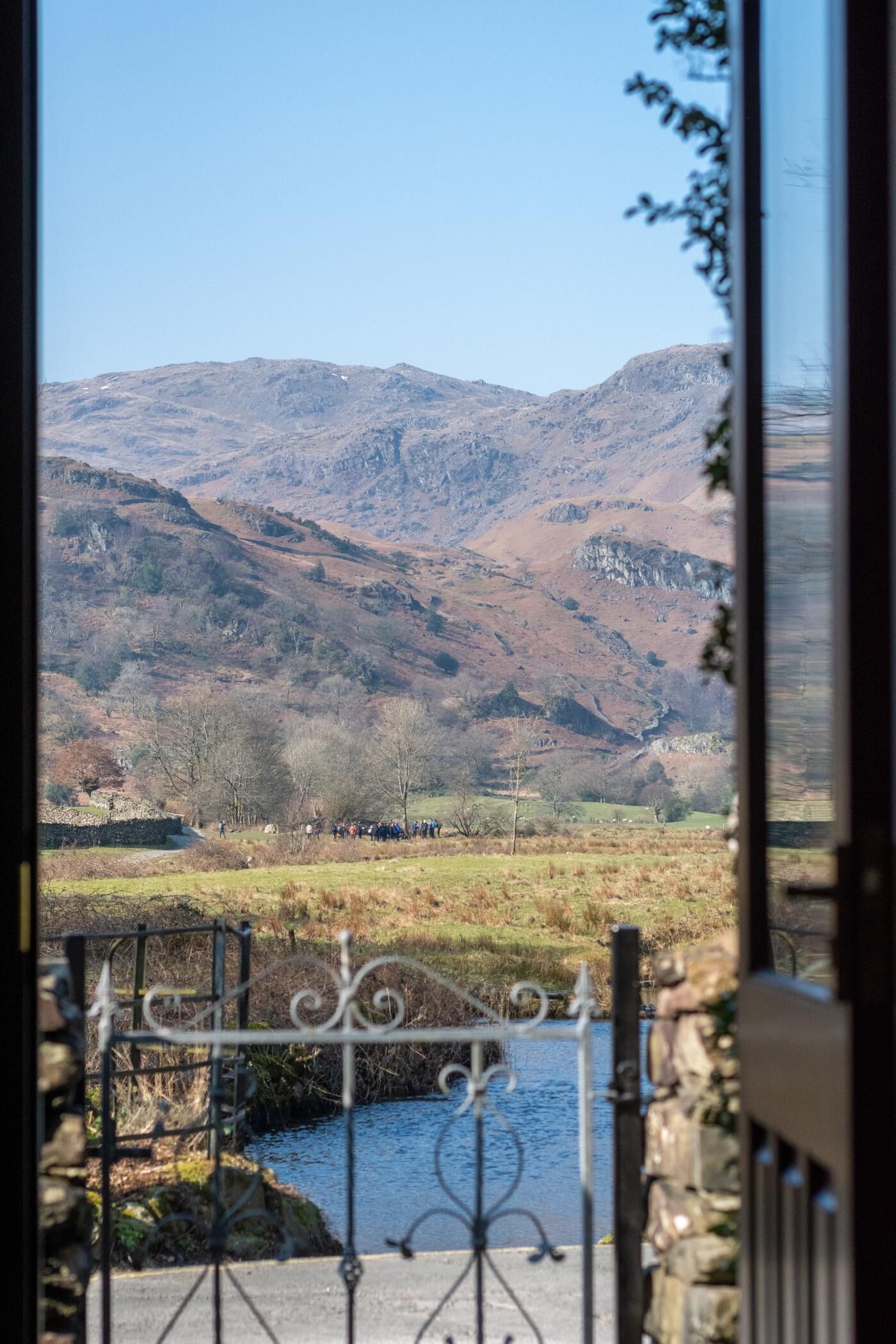 A view through a gate of a rural landscape with a river, stone wall, and mountains in the distance.