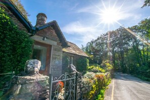 A stone cottage with a wooden door and a chimney, surrounded by lush greenery and a stone wall with flowers. - 2 Bed in Grasmere (oc-gl081) (Grasmere)