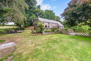 Une maison en pierre avec un toit en pente, entourée d’une pelouse bien entretenue et d’arbres.