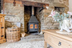Une cheminée en pierre avec un poêle à bois noir, un panier en osier et une commode en bois sur laquelle est posé un vase de fleurs.