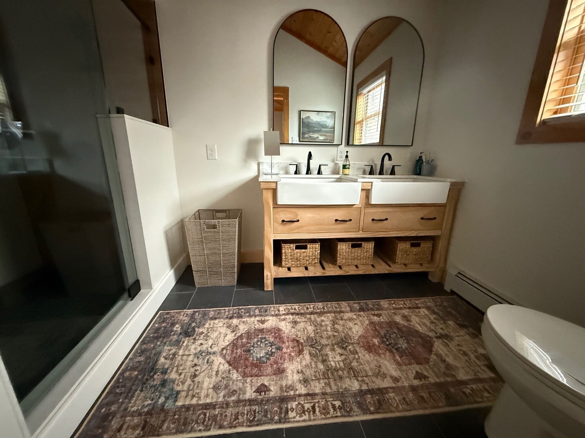 Une salle de bain dotée d’une douche à l’italienne, d’un meuble-lavabo double avec des tiroirs en bois, d’un grand miroir et d’un tapis à motifs sur le sol.